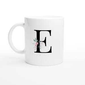 Könnte beinhalten: Weiße Keramiktasse mit dem schwarzen Buchstaben "E" und einem floralen Design in Rosa und Grün. Die Tasse hat einen gebogenen Henkel und eine glänzende Oberfläche. Das florale Design enthält kleine Sprenkel. Geeignet für heiße oder kalte Getränke.