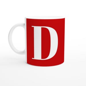 May include: White ceramic mug with a bright red background and a large white letter "D" printed on the front. The mug has a curved handle and a glossy finish, perfect for coffee or tea.