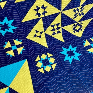 A quilt with a blue background and yellow and blue geometric patterns. The quilt is made of fabric squares sewn together to create a patchwork design and quilted with a repeated wavy line pantograph design.