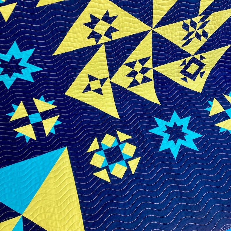 A quilt with a blue background and yellow and blue geometric patterns. The quilt is made of fabric squares sewn together to create a patchwork design and quilted with a repeated wavy line pantograph design.
