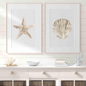 Può includere: Due stampe incorniciate ad acquerello con temi costieri. Una stampa mostra una stella marina, l'altra una conchiglia. Entrambe sono in tonalità neutre, su sfondo bianco e incorniciate in legno chiaro. L'opera d'arte è esposta su una cassettiera bianca.