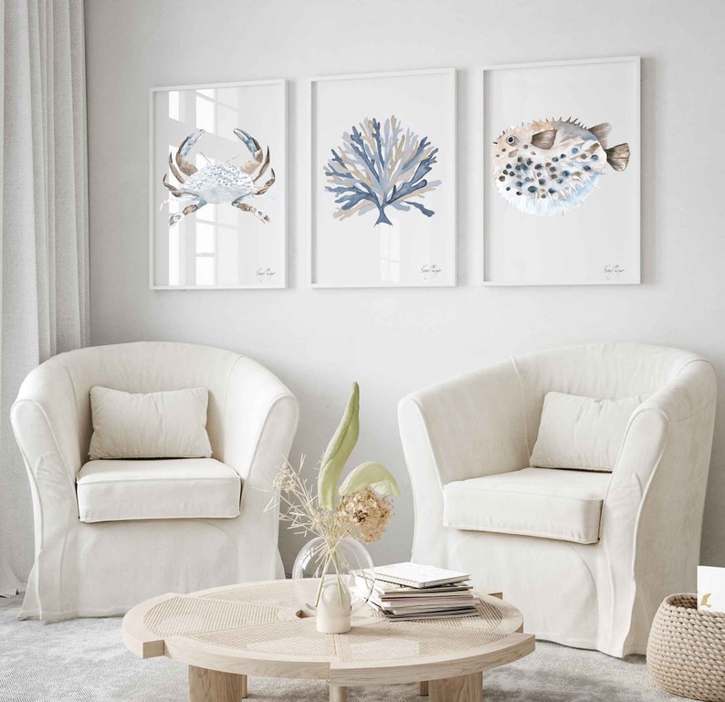 May include: Three framed watercolour prints of sea life, including a crab, coral, and a pufferfish. The prints are hung on a white wall above two white armchairs and a wooden coffee table.