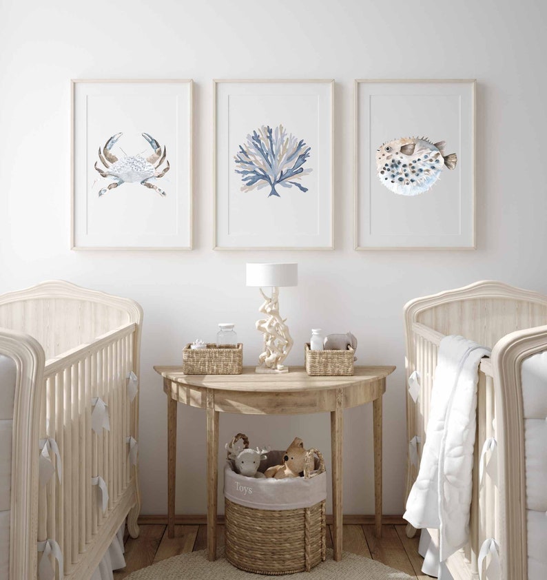May include: A nursery room with two cots, a wooden side table with a lamp, and three framed watercolour prints of a crab, coral, and a pufferfish. The table has a wicker basket with the word "Toys" on it.