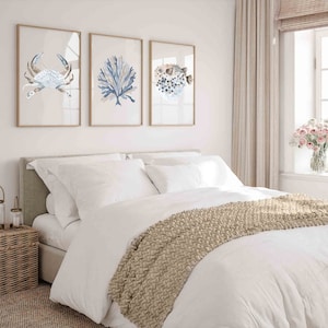 May include: A white bed with a beige knitted throw blanket. Three framed watercolour paintings of a crab, coral, and a pufferfish hang on the wall above the bed.