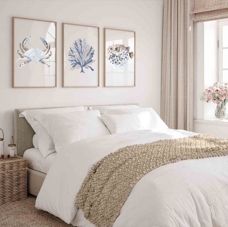 May include: A white bed with a beige knitted throw blanket. Three framed watercolour paintings of a crab, coral, and a pufferfish hang on the wall above the bed.