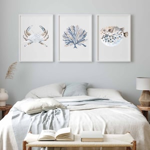 May include: Three framed watercolour prints of sea life, featuring a crab, coral, and a pufferfish. The prints are in shades of blue and brown and are displayed on a white wall above a bed with white bedding.