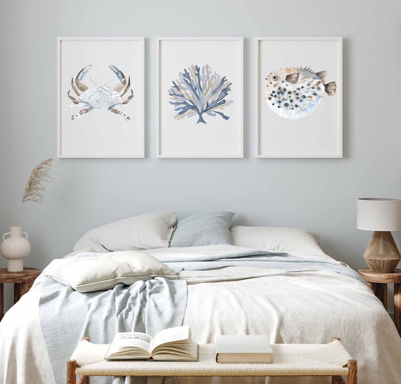 May include: Three framed watercolour prints of sea life, featuring a crab, coral, and a pufferfish. The prints are in shades of blue and brown and are displayed on a white wall above a bed with white bedding.