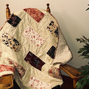 May include: A handmade quilt draped over a wooden chair. The quilt features a cream-colored background with rectangular patches of floral patterns in red, black, and cream. The chair is made of wood and has a curved back.