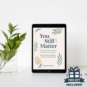 Könnte beinhalten: Ein schwarzes Tablet zeigt ein Journal-Cover mit dem Text "You Still Matter" und dem Untertitel "A Self-Care Reset Journal for Healthcare Workers". Das Cover-Design umfasst botanische Illustrationen und eine weiche beige Form. Eine Grafik "FREE GIFT INCLUDED" ist sichtbar.