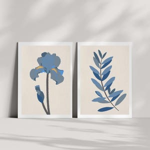 Könnte beinhalten: Zwei gerahmte Kunstdrucke mit botanischen Illustrationen. Ein Druck zeigt eine blaue Irisblüte mit gelben Akzenten und einem dunklen Stiel. Der andere zeigt einen blauen Blattzweig vor einem neutralen Hintergrund.