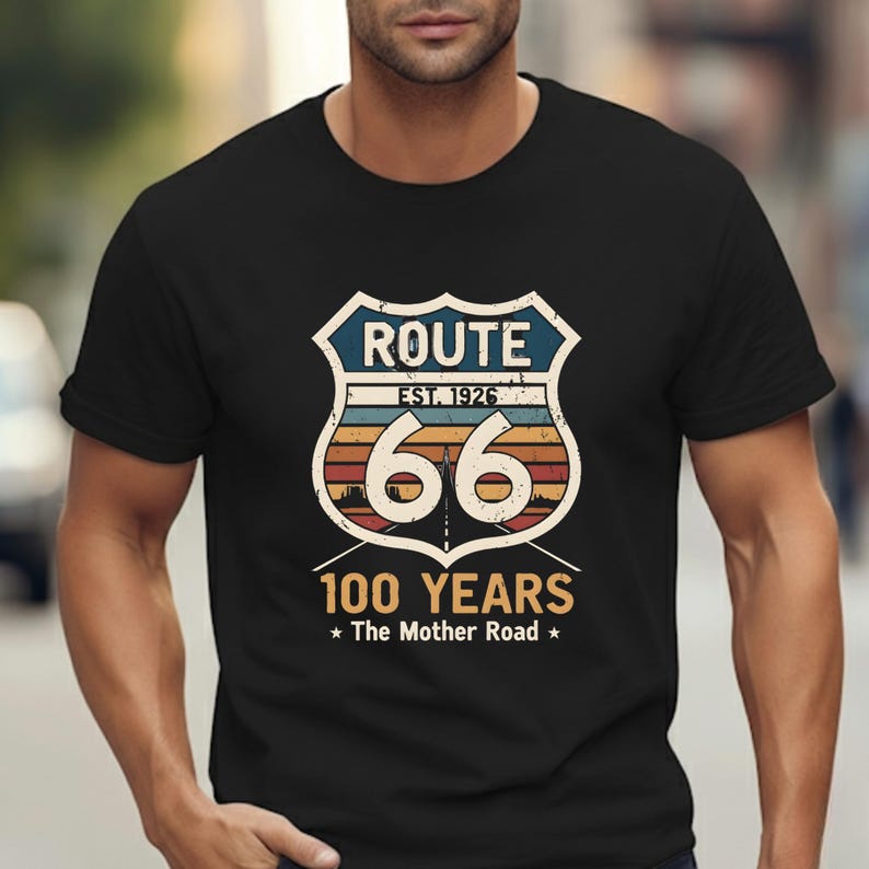 May include: A black short-sleeved t-shirt featuring a graphic of a Route 66 shield emblem with the text "ROUTE", "EST. 1926", "66", "100 YEARS", and "The Mother Road". The design includes a sunset-colored horizon line and a road perspective.