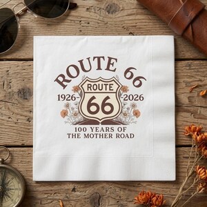 May include: A square white paper napkin featuring a brown graphic design of the Route 66 shield, the years 1926-2026, and the text 100 YEARS OF THE MOTHER ROAD. The design includes illustrations of wildflowers surrounding the central shield emblem.