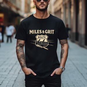 May include: Black t-shirt featuring a beige Route 66 graphic and the words "MILES & GRIT". The graphic includes the Route 66 shield with the number 66. The shirt is worn by a person with tattoos and sunglasses, standing in a street.