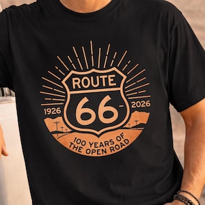 Könnte beinhalten: Schwarzes T-Shirt mit einem Route 66-Design in Orange und Beige. Die Grafik enthält den Text "ROUTE 66" über der Nummer "66", mit den Jahren "1926" und "2026" auf beiden Seiten. Der Satz "100 YEARS OF THE OPEN ROAD" steht unten.