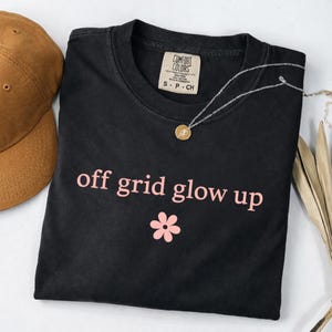 May include: Black t-shirt with the text "off grid glow up" in pink, along with a pink flower graphic. A brown baseball cap and a silver necklace with a pendant are also visible.