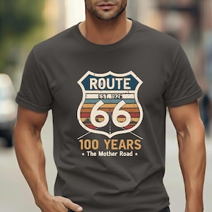 May include: A charcoal gray short-sleeve t-shirt featuring a graphic of a Route 66 shield emblem. The design includes the text "ROUTE", "EST. 1926", "66", "100 YEARS", and "The Mother Road" with a sunset-colored horizon and a road perspective.