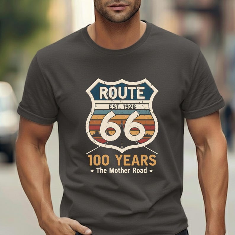 May include: A charcoal gray short-sleeve t-shirt featuring a graphic of a Route 66 shield emblem. The design includes the text "ROUTE", "EST. 1926", "66", "100 YEARS", and "The Mother Road" with a sunset-colored horizon and a road perspective.