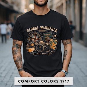 May include: Black t-shirt with a travel-themed graphic. The design includes a compass, camera, passport, map, and film reel, with the words "GLOBAL WANDERER" and "EST. 2024". The bottom of the shirt has the text "COMFORT COLORS 1717".