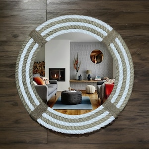 Op de afbeelding: Ronde spiegel met een touwframe. Het frame heeft afwisselende banden van wit en beige touw, vastgezet met touw. De spiegel reflecteert een woonkamer met een open haard en meubels.