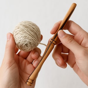 Pode incluir: Um fuso de madeira e uma bola de fio de cor natural. O fuso é castanho claro com detalhes em castanho escuro. O fio é de cor bege claro. Os itens são segurados em duas mãos contra um fundo branco.