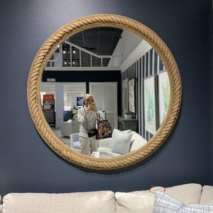 Op de afbeelding: Een ronde spiegel met een dik, touwachtig frame hangt aan een donkerblauwe muur. De spiegel reflecteert een kamer met witte meubels en kunstwerken. Het frame is lichtbruin van kleur en voegt een nautische touch toe.