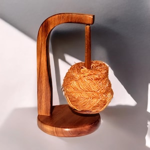 May include: A wooden yarn swift with a ball of orange yarn. The swift is made of polished wood and has a curved arm and a round base. The yarn is a bright orange color and is wound into a ball.