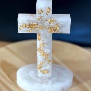 May include: A white, semi-translucent cross sculpture featuring embedded gold leaf flakes throughout the structure. The cross is mounted vertically on a matching circular white base, creating a stable, upright decorative object with a smooth finish.