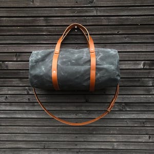 May include: A dark grey canvas duffel bag with brown leather straps and buckles. The bag is hanging from a wooden wall.