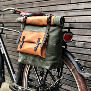 Könnte beinhalten: Eine grüne Canvas- und braune Leder-Fahrradtasche mit verstellbaren Riemen, die an einem schwarzen Fahrrad befestigt ist.