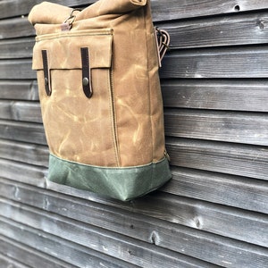 May include: A brown canvas backpack with a roll-top closure and a green bottom. The backpack has a large front pocket with a leather flap and buckle.
