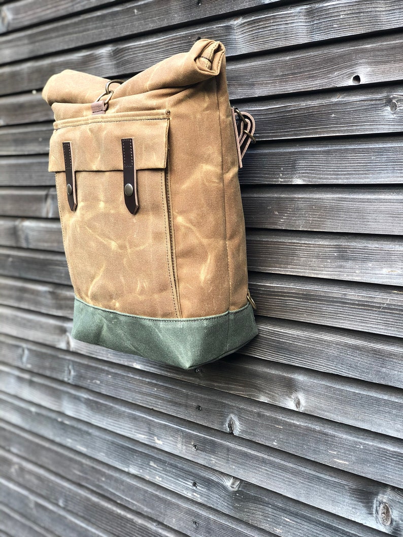 May include: A brown canvas backpack with a roll-top closure and a green bottom. The backpack has a large front pocket with a leather flap and buckle.