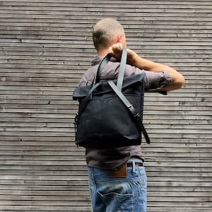 May include: A person wearing a dark grey shirt and blue jeans is adjusting a black canvas backpack with leather straps.