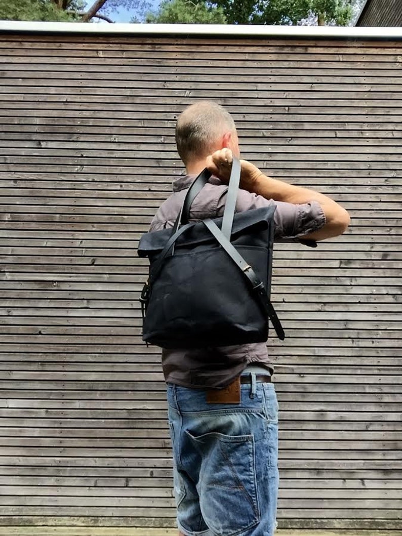 May include: A person wearing a dark grey shirt and blue jeans is adjusting a black canvas backpack with leather straps.