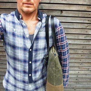 May include: A man wearing a blue and gray plaid shirt with a black and brown canvas tote bag slung over his shoulder.