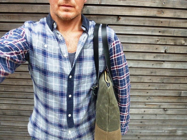 May include: A man wearing a blue and gray plaid shirt with a black and brown canvas tote bag slung over his shoulder.