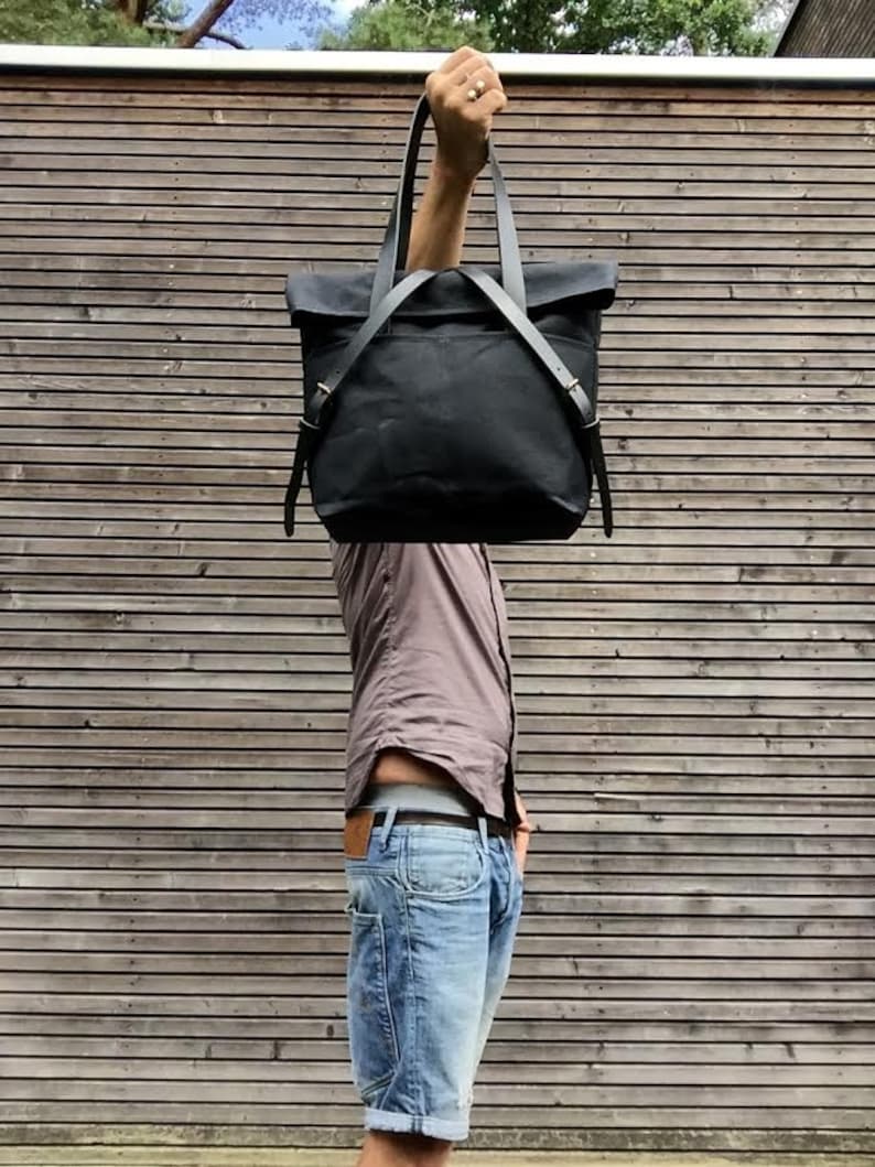 May include: A black canvas tote bag with leather straps and buckles. The bag is being held up by a person's hand.
