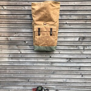 May include: A brown canvas backpack with leather straps and a green bottom. The backpack is hanging on a wooden wall.
