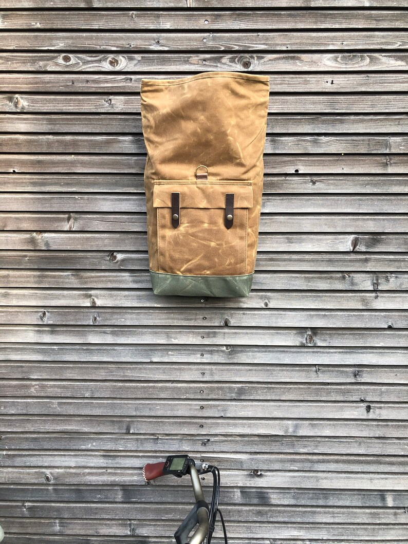 May include: A brown canvas backpack with leather straps and a green bottom. The backpack is hanging on a wooden wall.