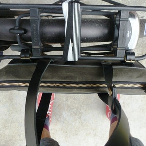 May include: A green canvas tote bag with black leather straps and a gold zipper. The bag is attached to a black bike rack with a black and white elastic strap.