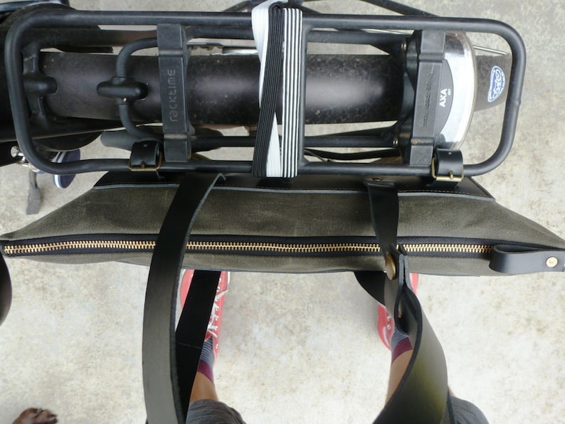 May include: A green canvas tote bag with black leather straps and a gold zipper. The bag is attached to a black bike rack with a black and white elastic strap.