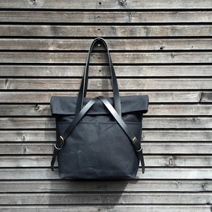 May include: A black canvas tote bag with leather straps and brass hardware. The bag is hanging on a wooden wall.