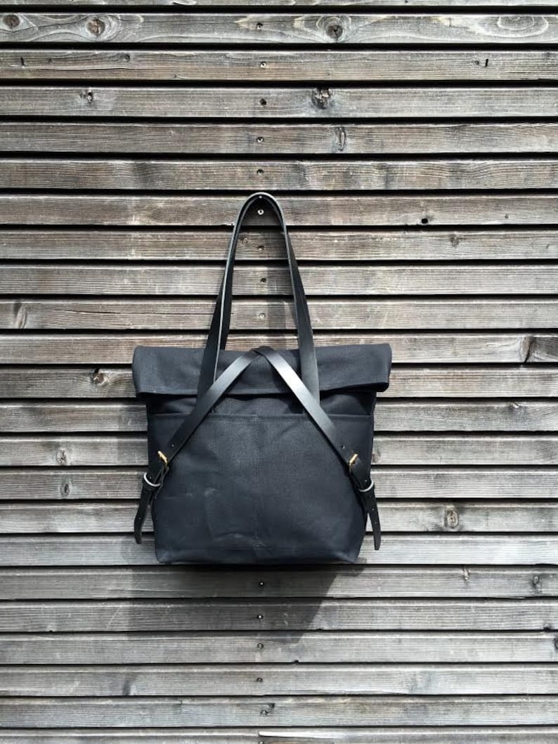 May include: A black canvas tote bag with leather straps and brass hardware. The bag is hanging on a wooden wall.