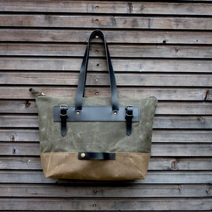 May include: A green and brown canvas tote bag with black leather handles and straps. The bag has two brass buckles and a brass button.