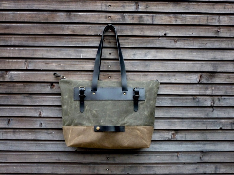 May include: A green and brown canvas tote bag with black leather handles and straps. The bag has two brass buckles and a brass button.