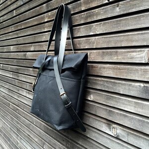 May include: A black canvas tote bag with a long, adjustable leather strap. The bag is hanging on a wooden wall.