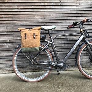 May include: A black electric bicycle with a brown canvas pannier bag attached to the rear rack. The bike has a black frame, black wheels, and a black seat. The bike is parked in front of a wooden wall.