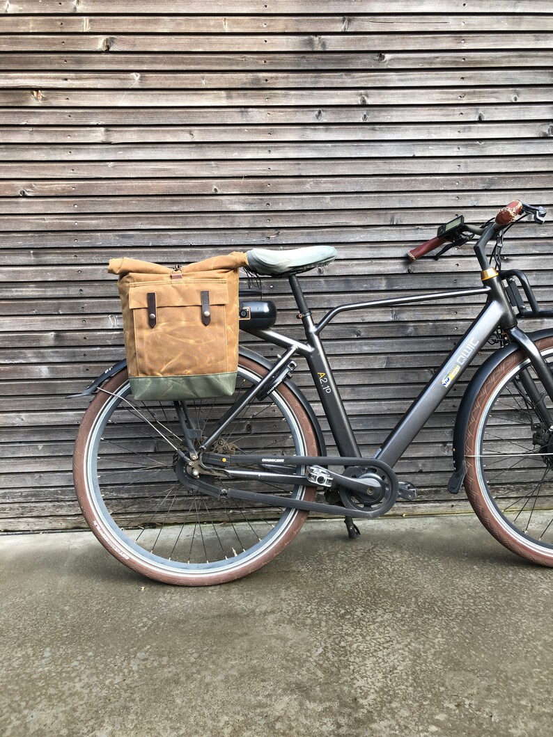 May include: A black electric bicycle with a brown canvas pannier bag attached to the rear rack. The bike has a black frame, black wheels, and a black seat. The bike is parked in front of a wooden wall.
