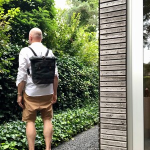 May include: A person wearing a white shirt and brown shorts is walking down a path. They are carrying a black canvas backpack with leather straps and buckles.