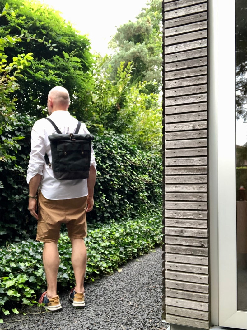 May include: A person wearing a white shirt and brown shorts is walking down a path. They are carrying a black canvas backpack with leather straps and buckles.