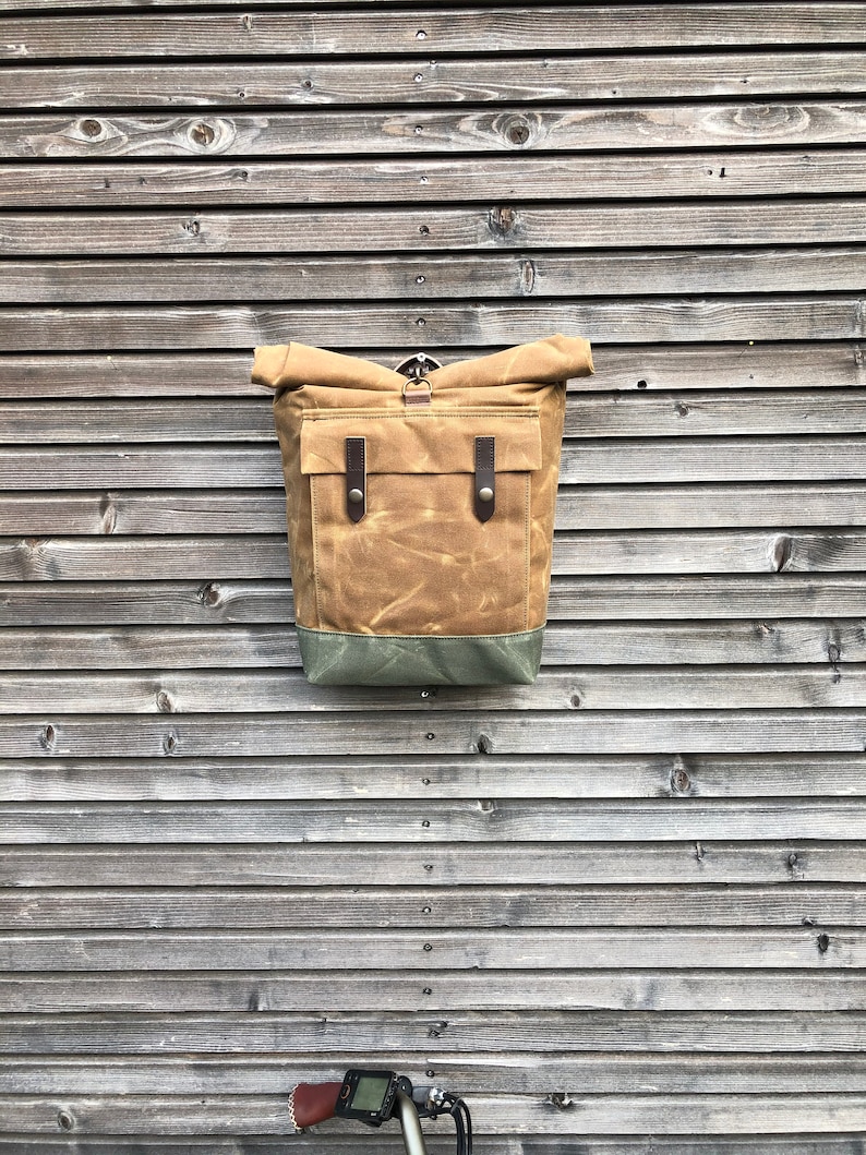 May include: A brown canvas backpack with a roll-top closure and leather accents. The backpack is hanging on a wooden wall.